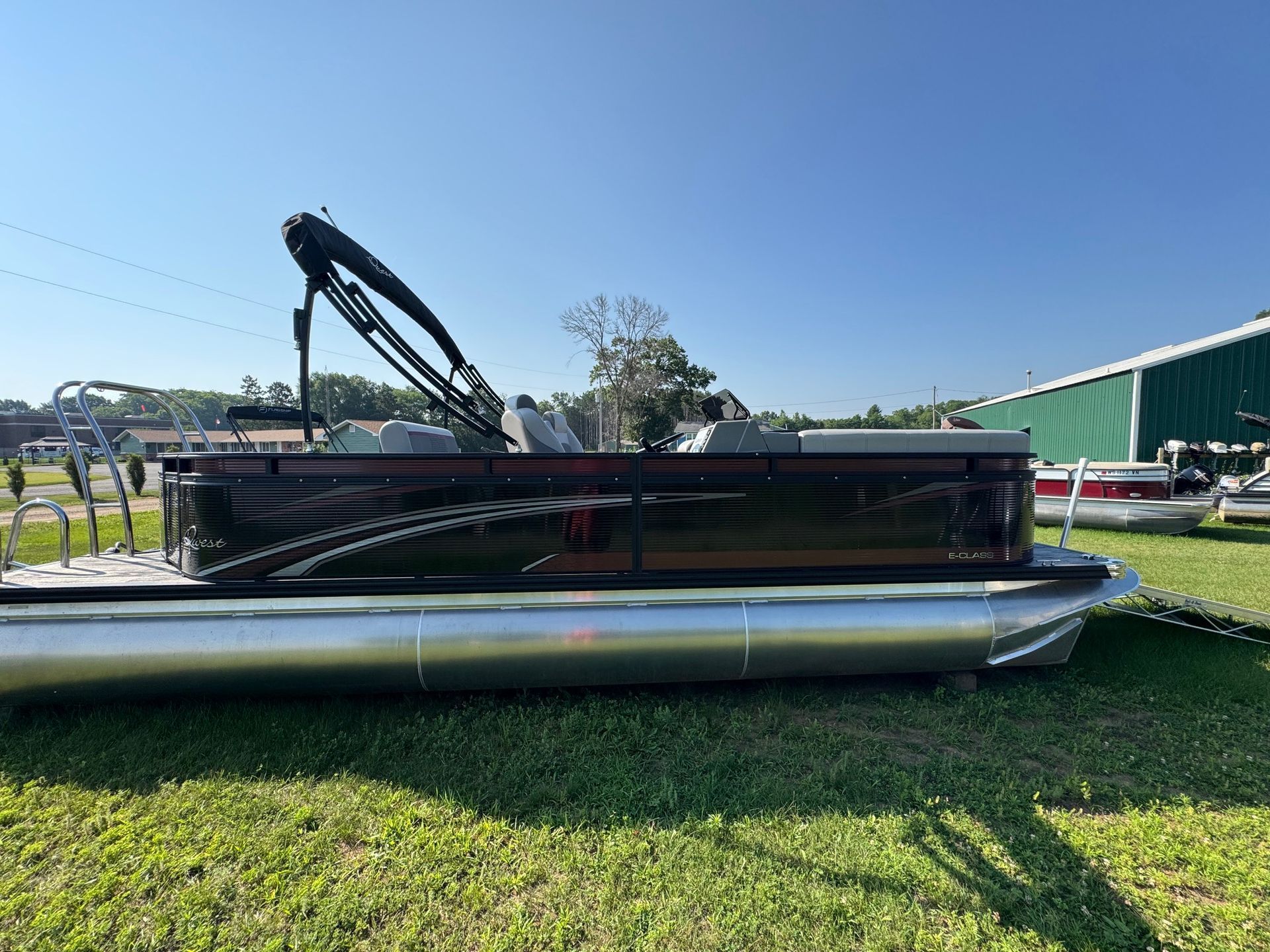 Pontoon boat on grass, under blue sky; black exterior, silver pontoons. Green and white building in the background.