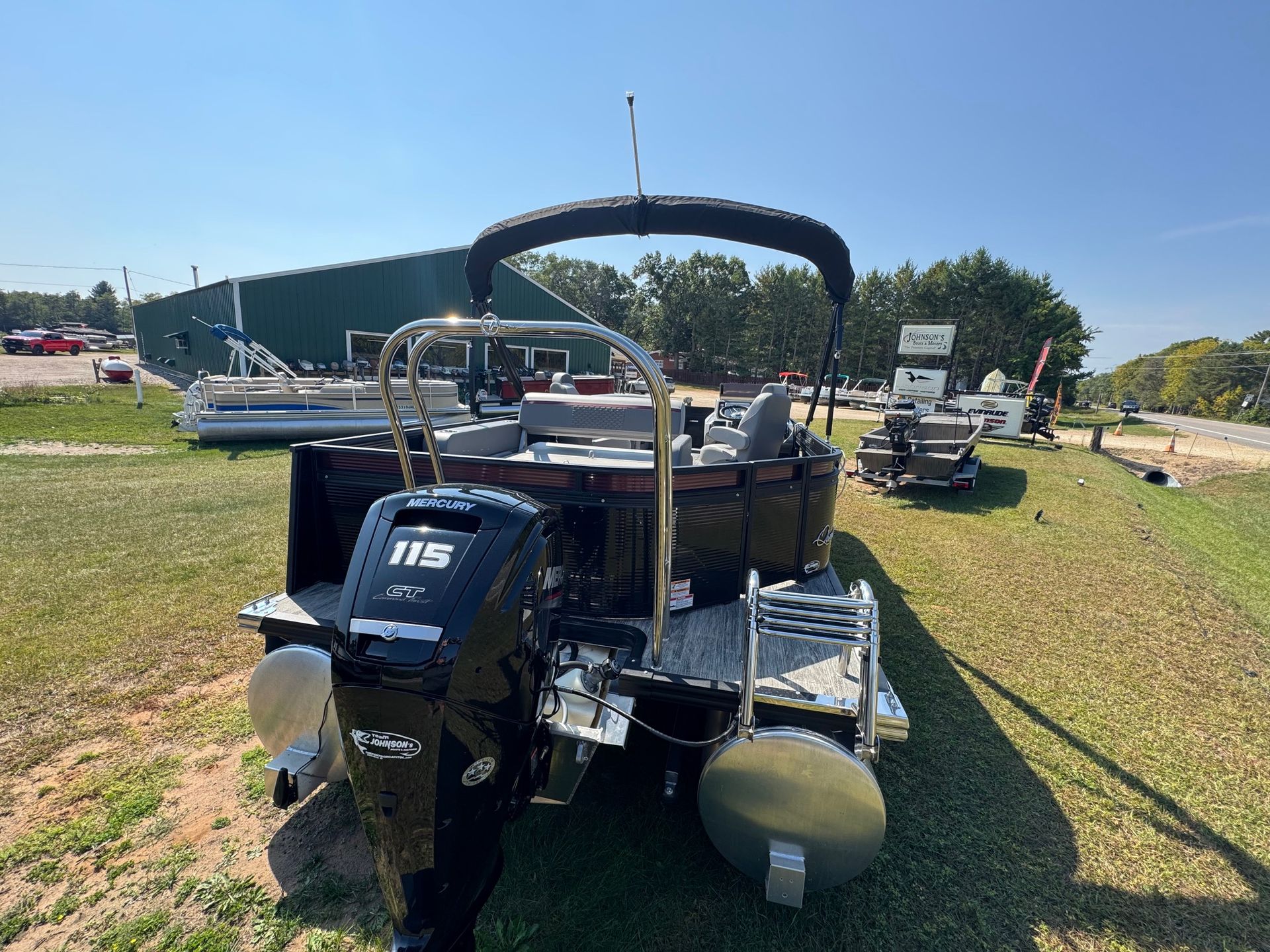 Black pontoon boat with a 115 HP motor, sitting on grass, under a blue sky.