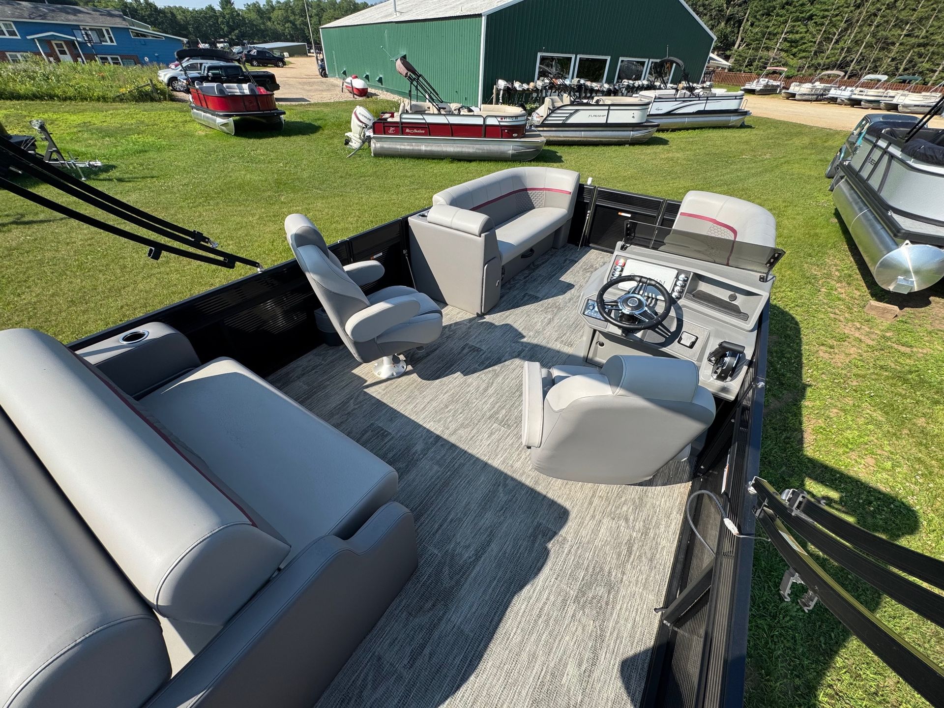 Pontoon boat interior with gray seating and steering wheel. Several other boats are in the background on the grass.