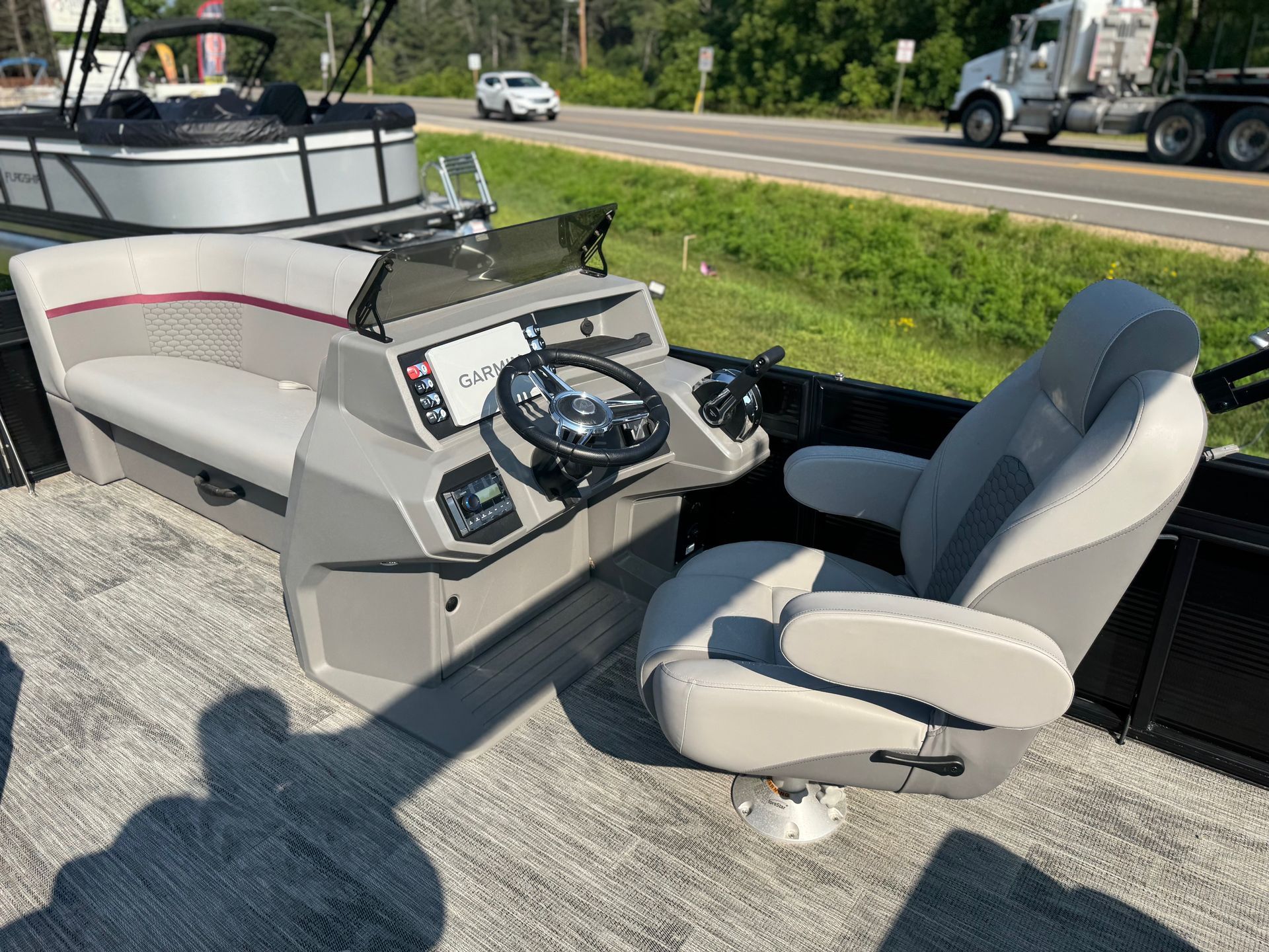 Gray pontoon boat helm with steering wheel, captain's chair, and bench seating. Road in background.