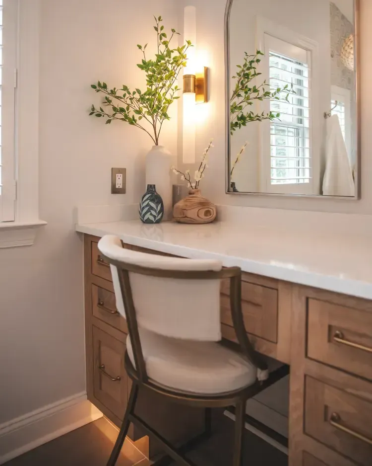 Bright vanity desk with chair, white countertop, wall mirror, and a lamp with flowers in a softly lit room