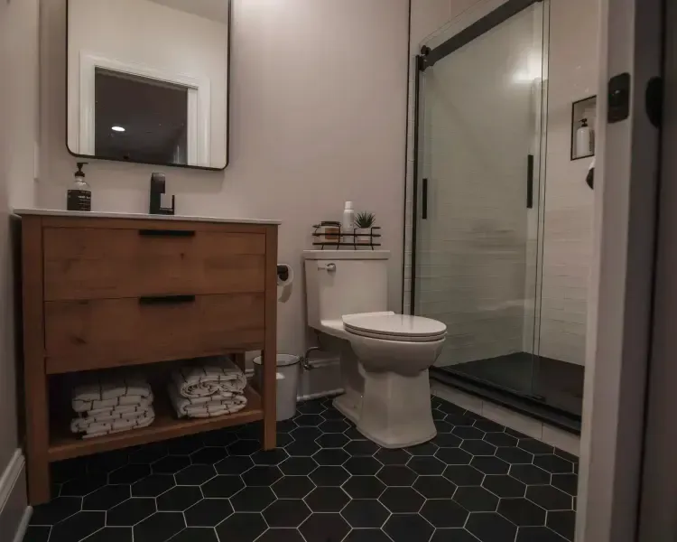 Modern bathroom with dark hex tile floor, wooden vanity, toilet, glass shower, and wall mirror