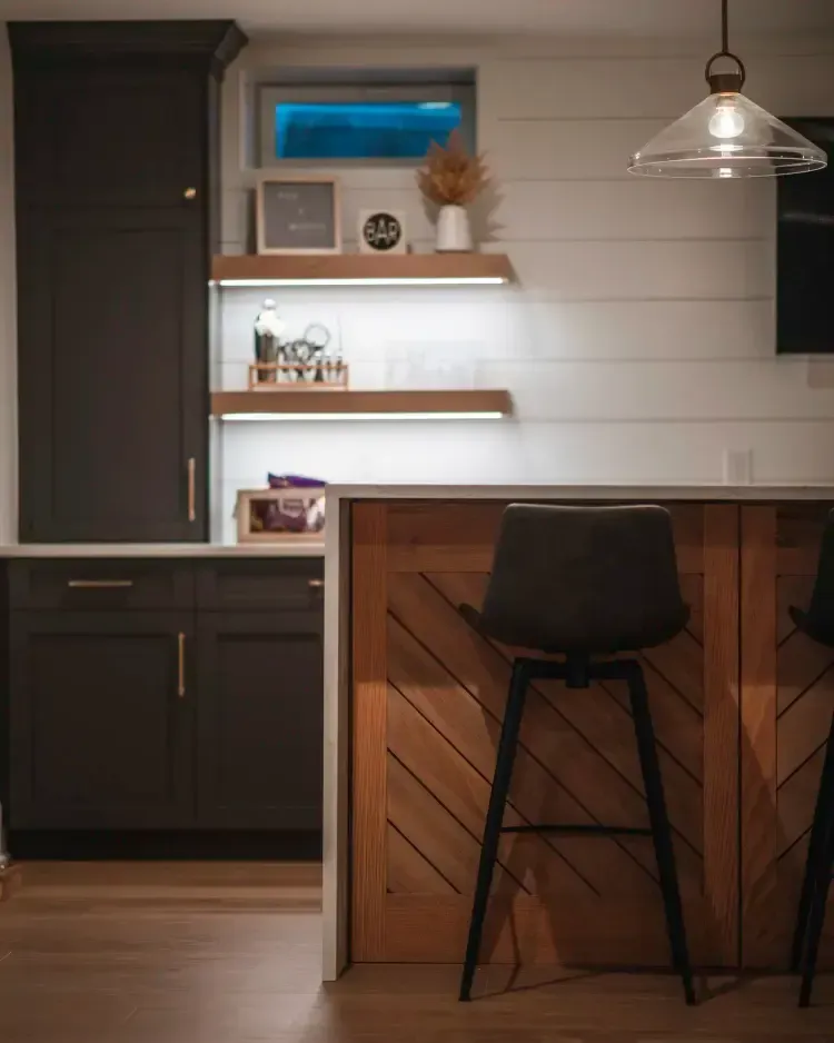Modern kitchen with dark cabinets, wood island, floating shelves, and a stool under pendant lighting.