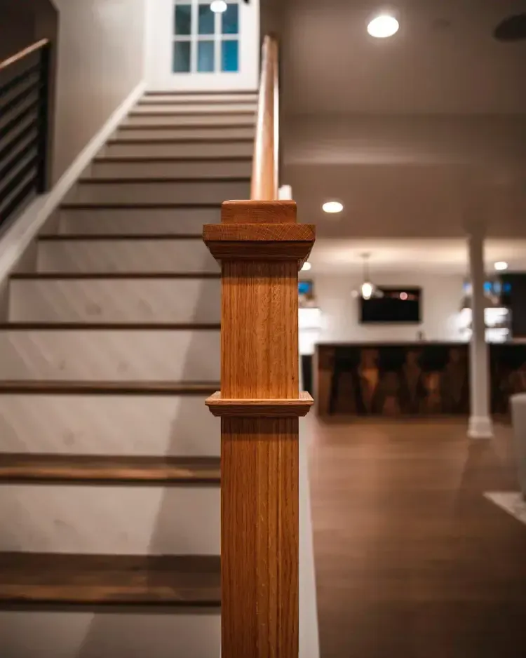Wooden staircase railing in a warmly lit interior hallway with stairs and a blurred room beyond