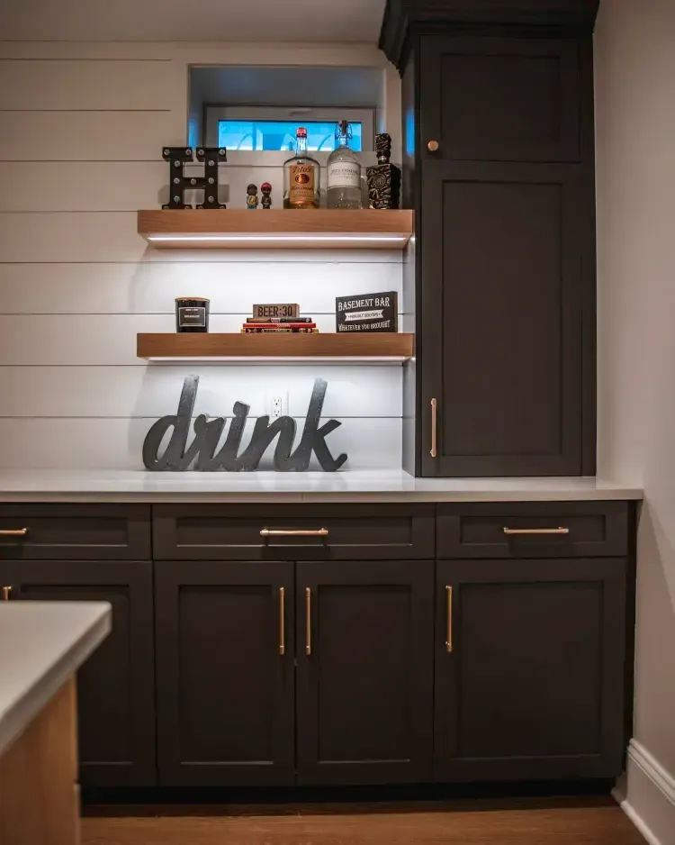 Dark kitchen wet bar with shelves, bottles, and a decorative “drink” sign above cabinets.