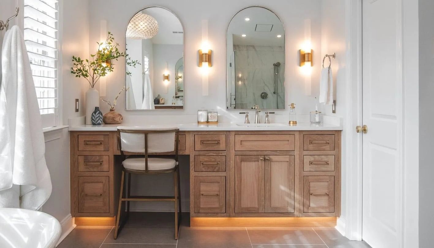 Bright vanity with three arched mirrors, warm sconces, and a stool in a white bathroom