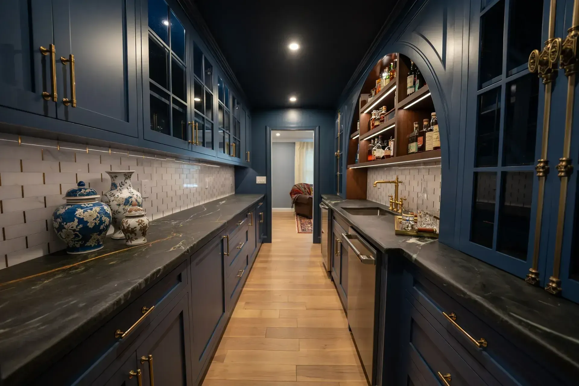 Narrow blue galley kitchen with dark cabinets, marble counters, wood floor, and warm overhead lights