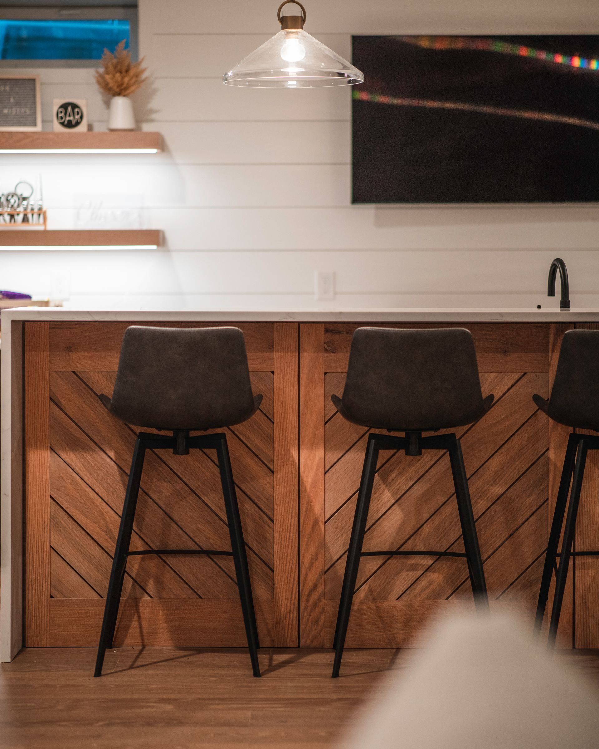 Modern kitchen bar with wood cabinets, white shiplap wall, pendant light, and black bar stools