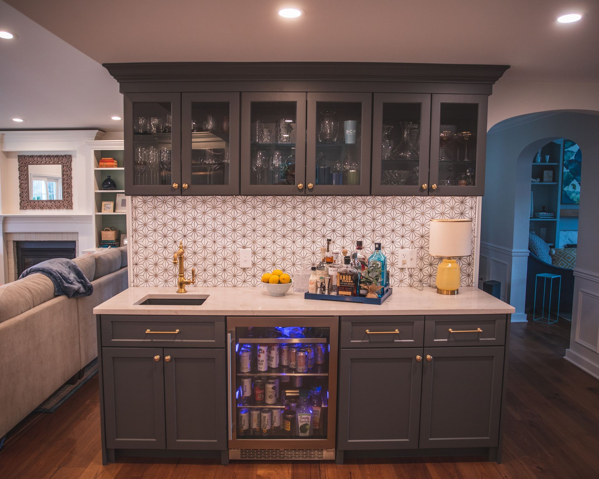 Dark gray kitchen island with glass-front cabinets, patterned backsplash, and lit beverage fridge in a modern home