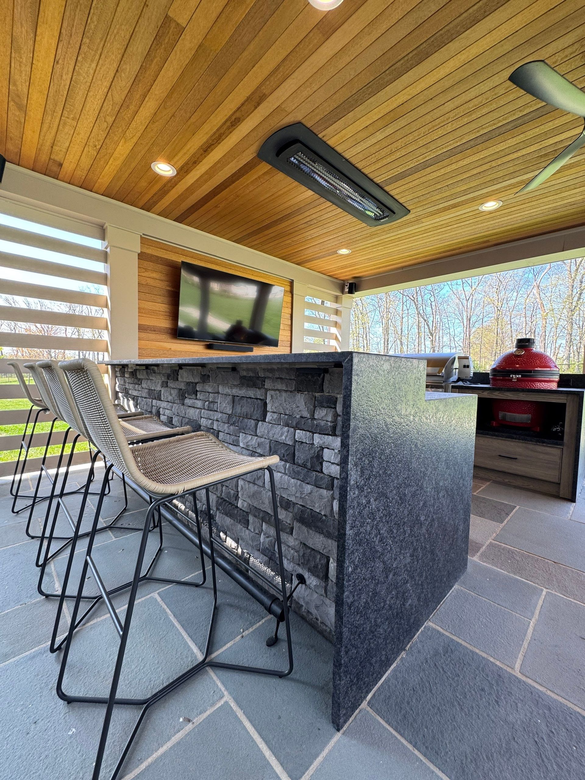 Covered outdoor bar with stools, wall-mounted TV, and a grill under a wood-paneled roof