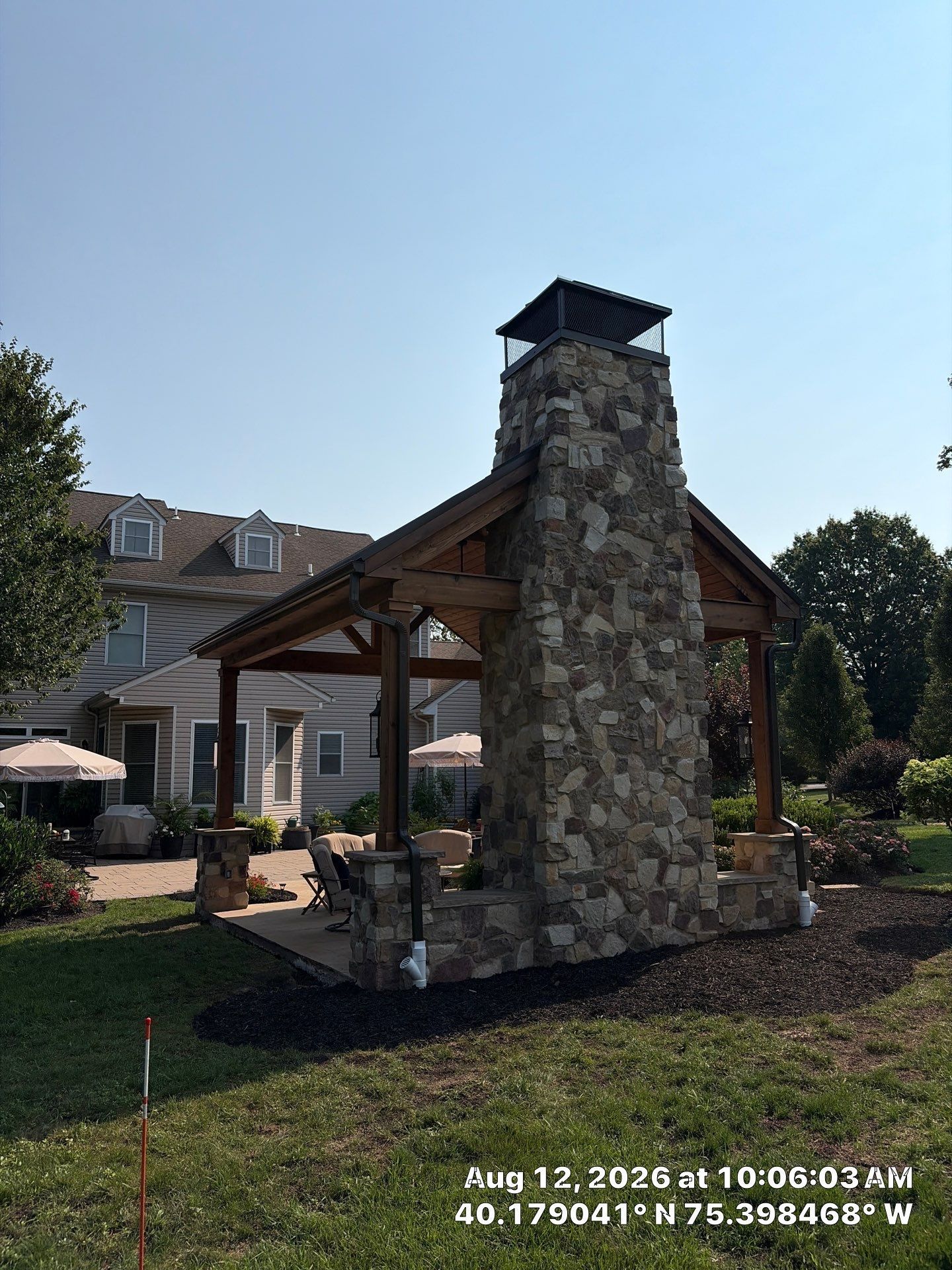 Stone outdoor fireplace under a wooden pavilion in a landscaped courtyard beside a house