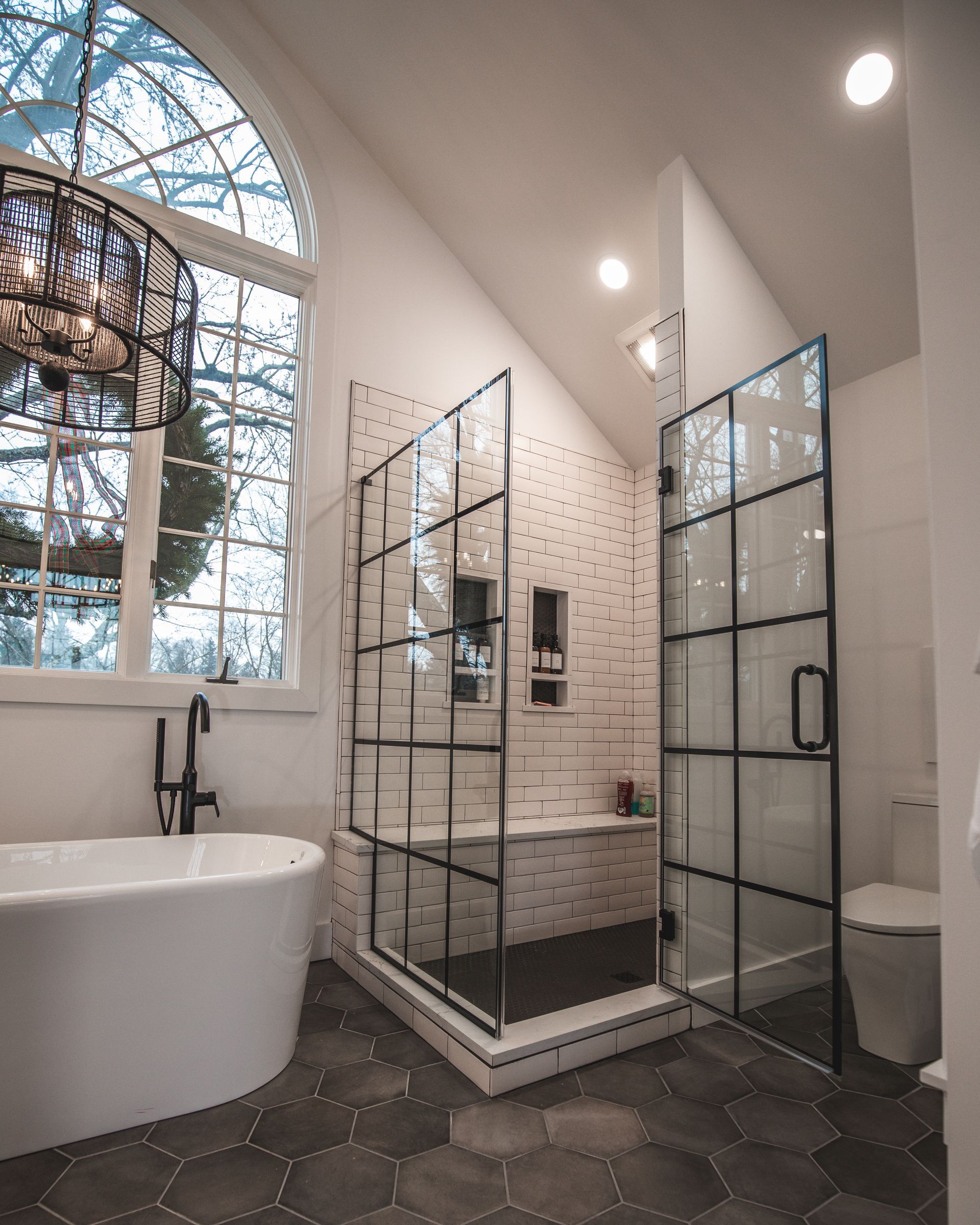 Modern bathroom with a glass shower, freestanding tub, hex tile floor, and large arched window