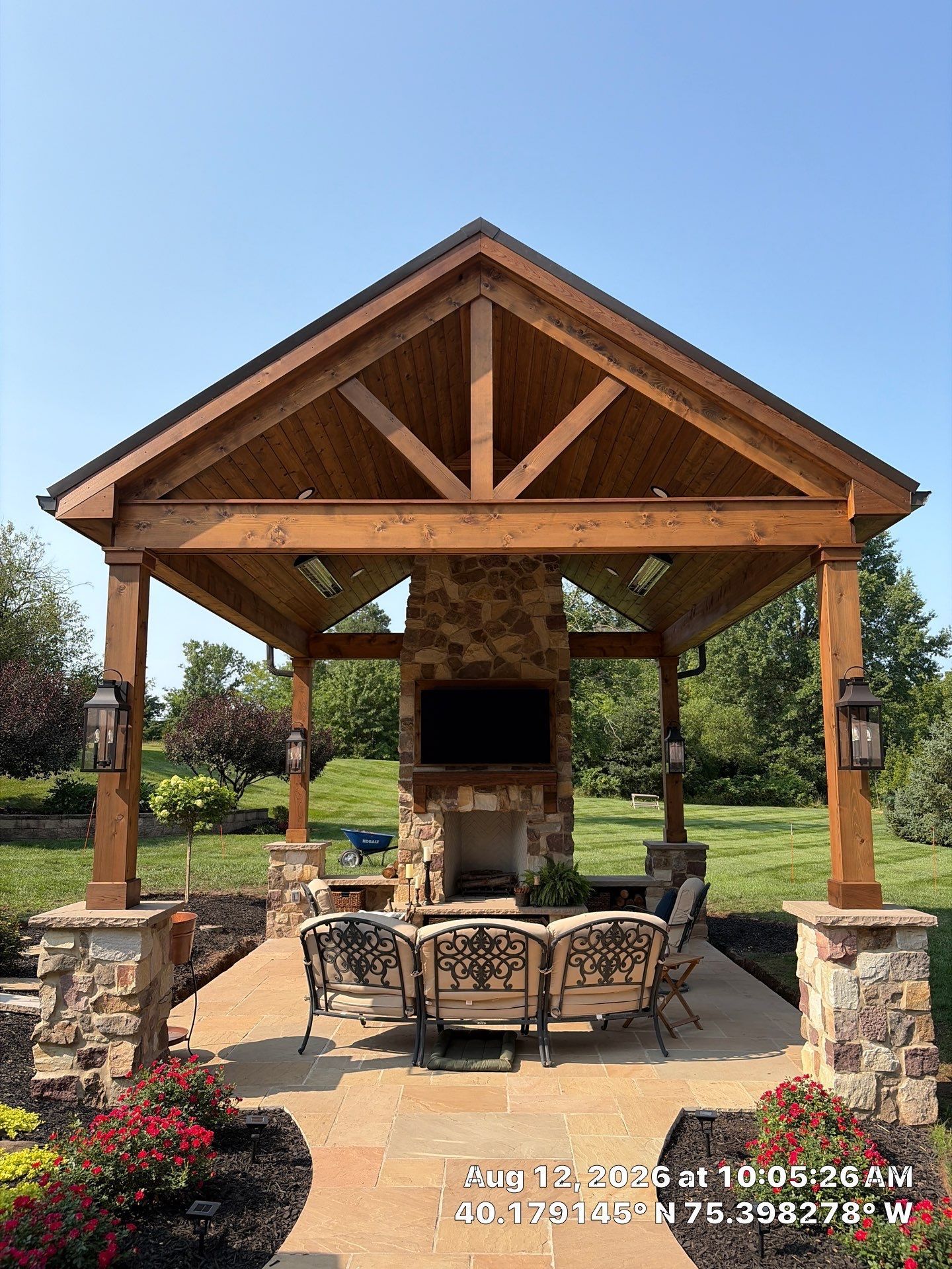 Stone outdoor pavilion with wooden roof, fireplace, and dining table in a landscaped garden