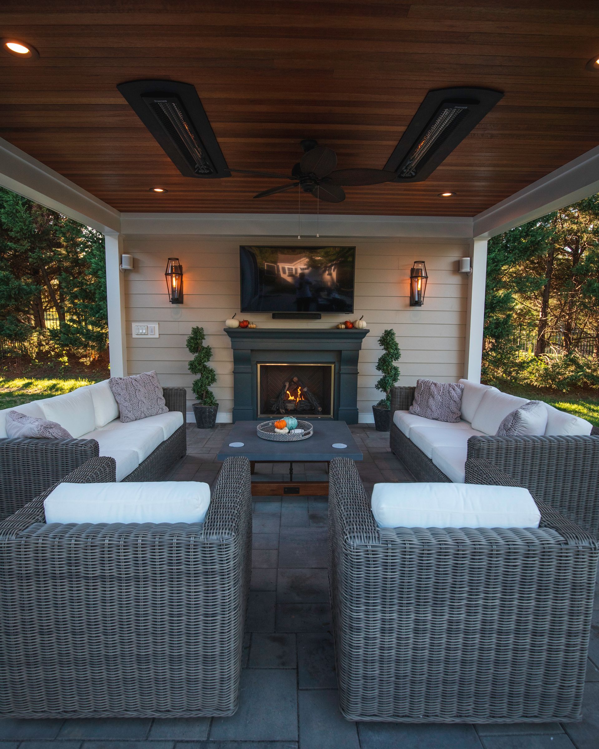 Covered patio lounge with wicker seating, fireplace, TV, and a wood ceiling overlooking a lush garden.
