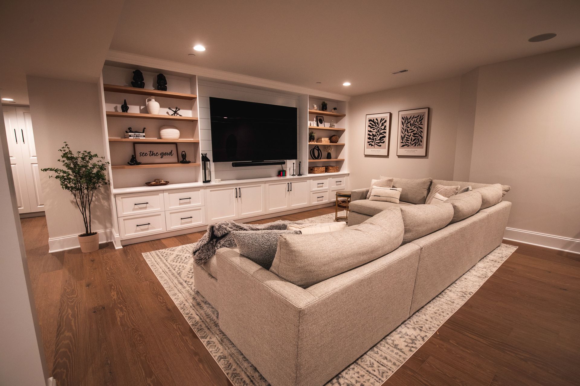 Bright living room with beige sectional sofa, wall-mounted TV, built-in shelves, and warm wood floors