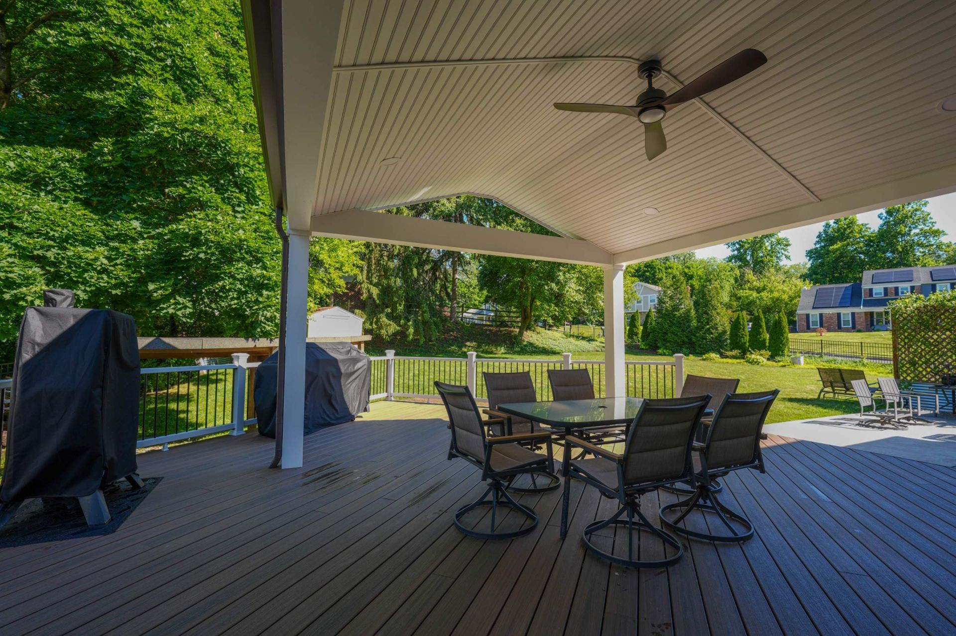 Covered backyard deck with outdoor dining table, chairs, grill, and green trees beyond the railing.