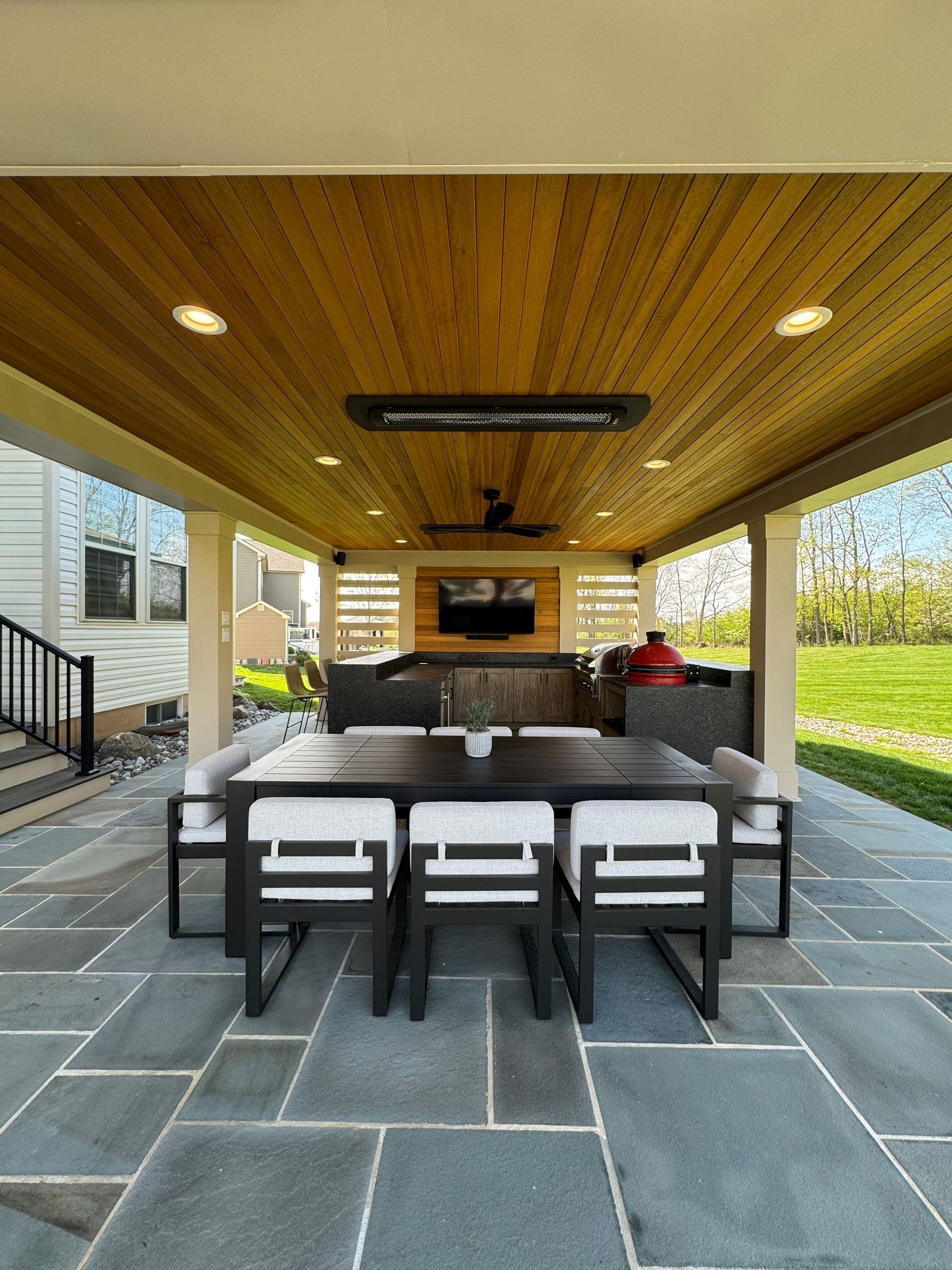 Covered patio with outdoor dining table, black chairs, grill, stone floor, and wood ceiling overlooking a grassy yard