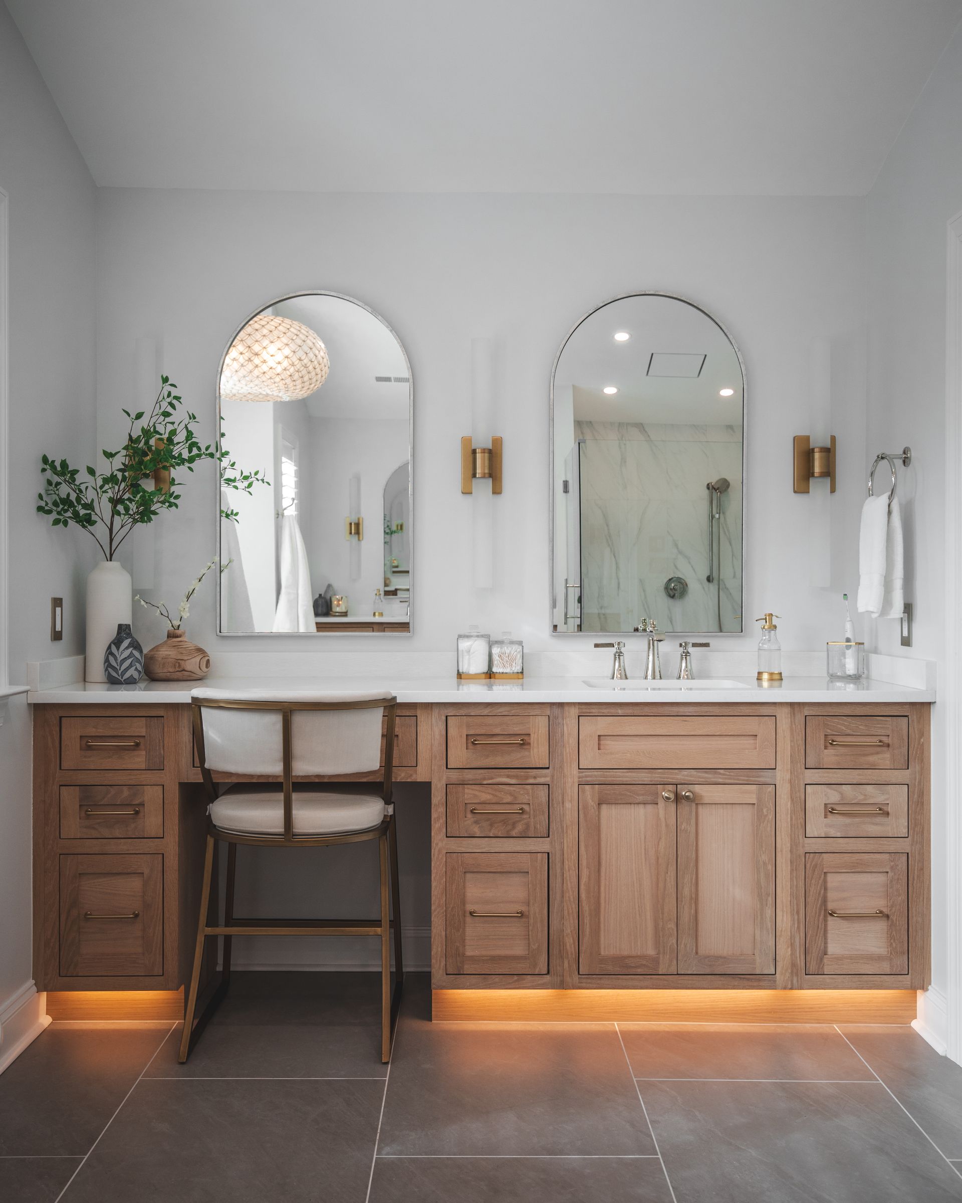 Modern bathroom vanity with double arched mirrors, wood cabinets, and a chair under soft lighting