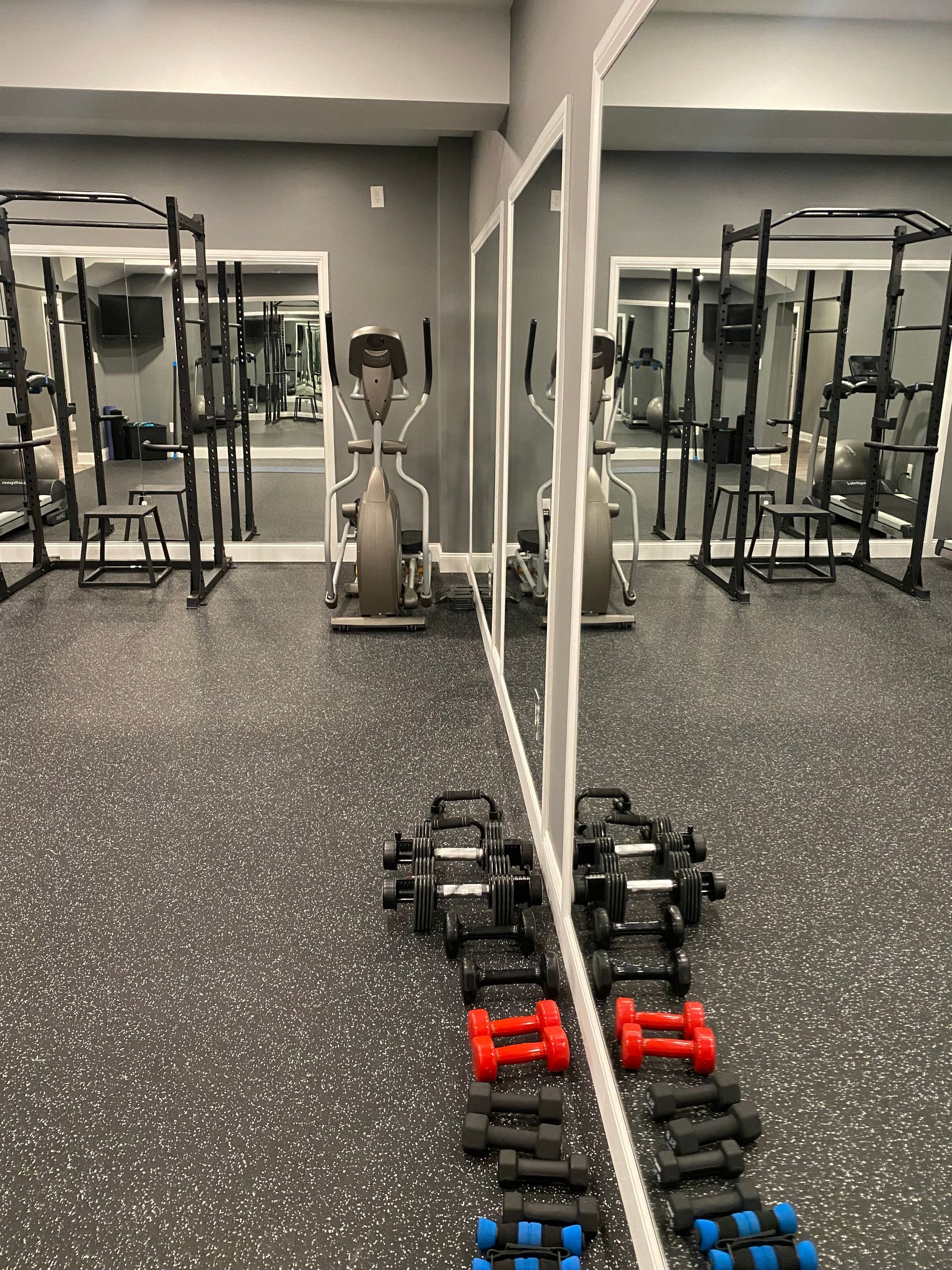 Gym with dumbbells and weight racks reflected in a large mirror on a speckled floor.