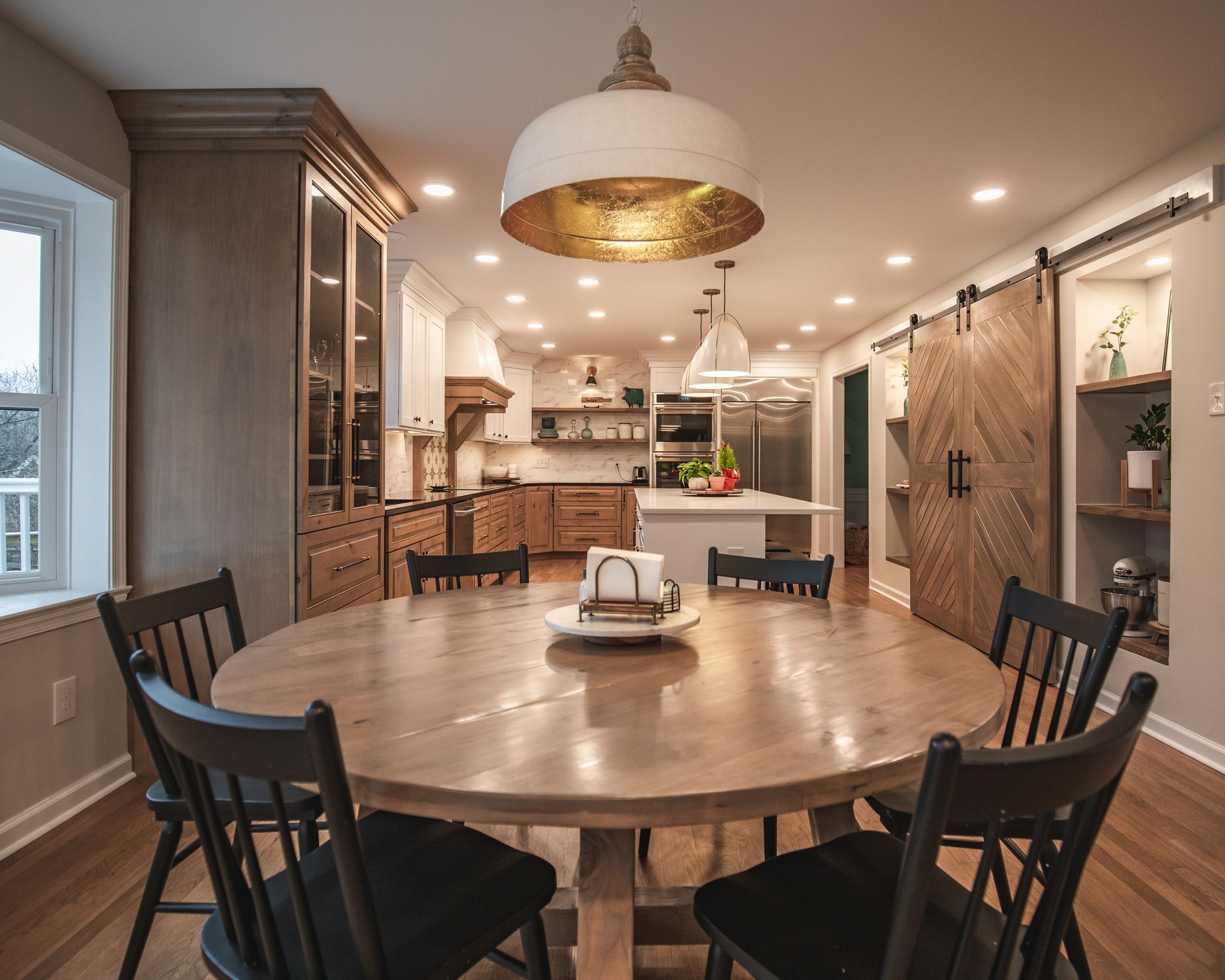 Bright dining room with a round wooden table, black chairs, and open kitchen in the background
