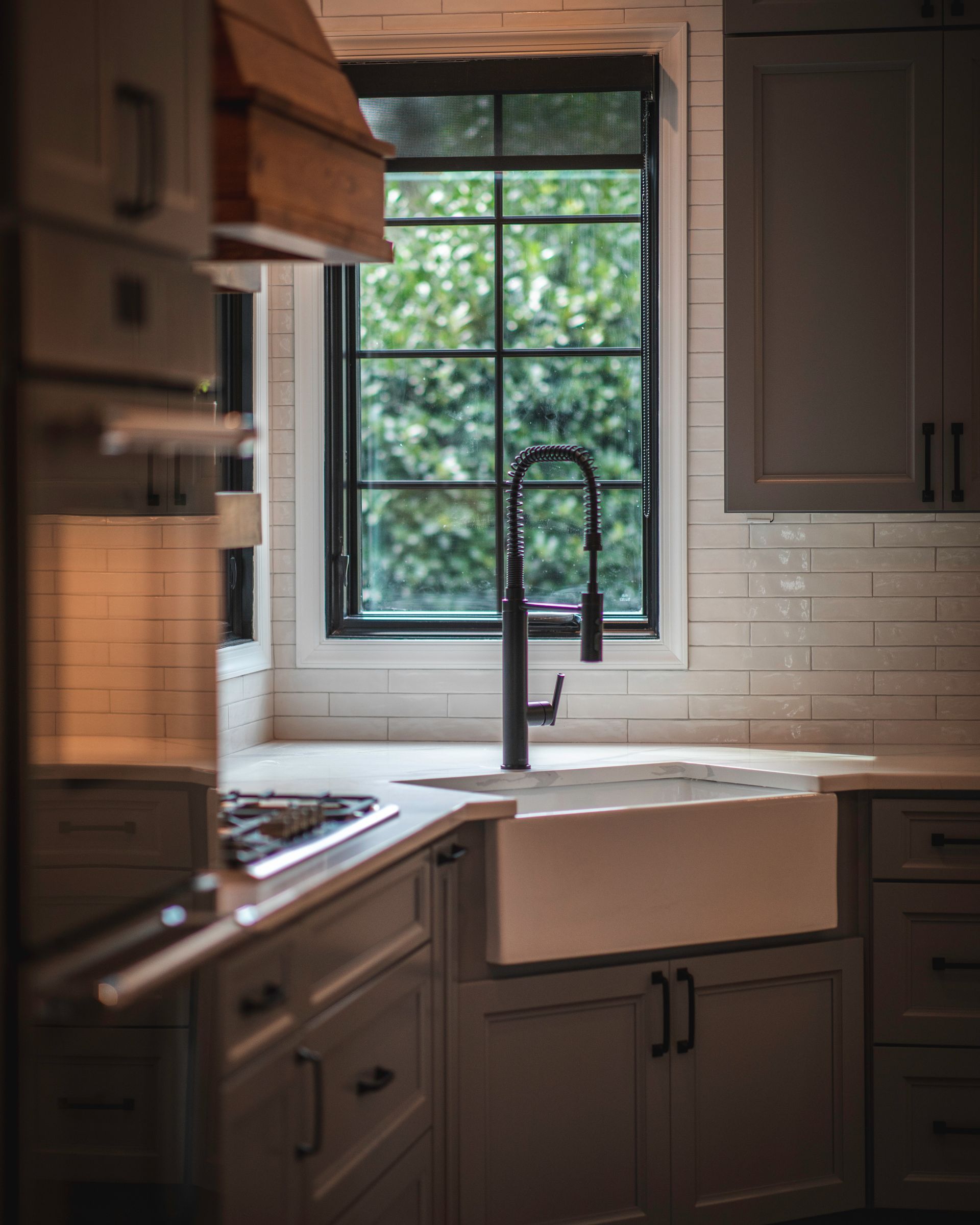 Sunlit kitchen sink beneath a window, with gray cabinets and white tile backsplash