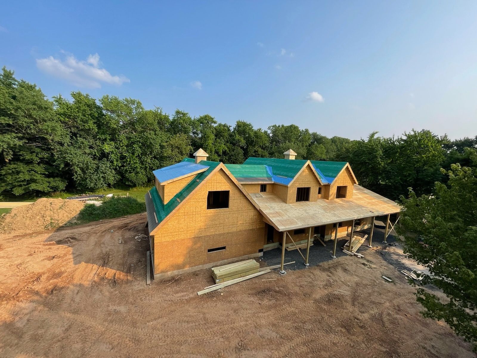 Partially built house with teal roof in a clearing beside dense green trees under a blue sky