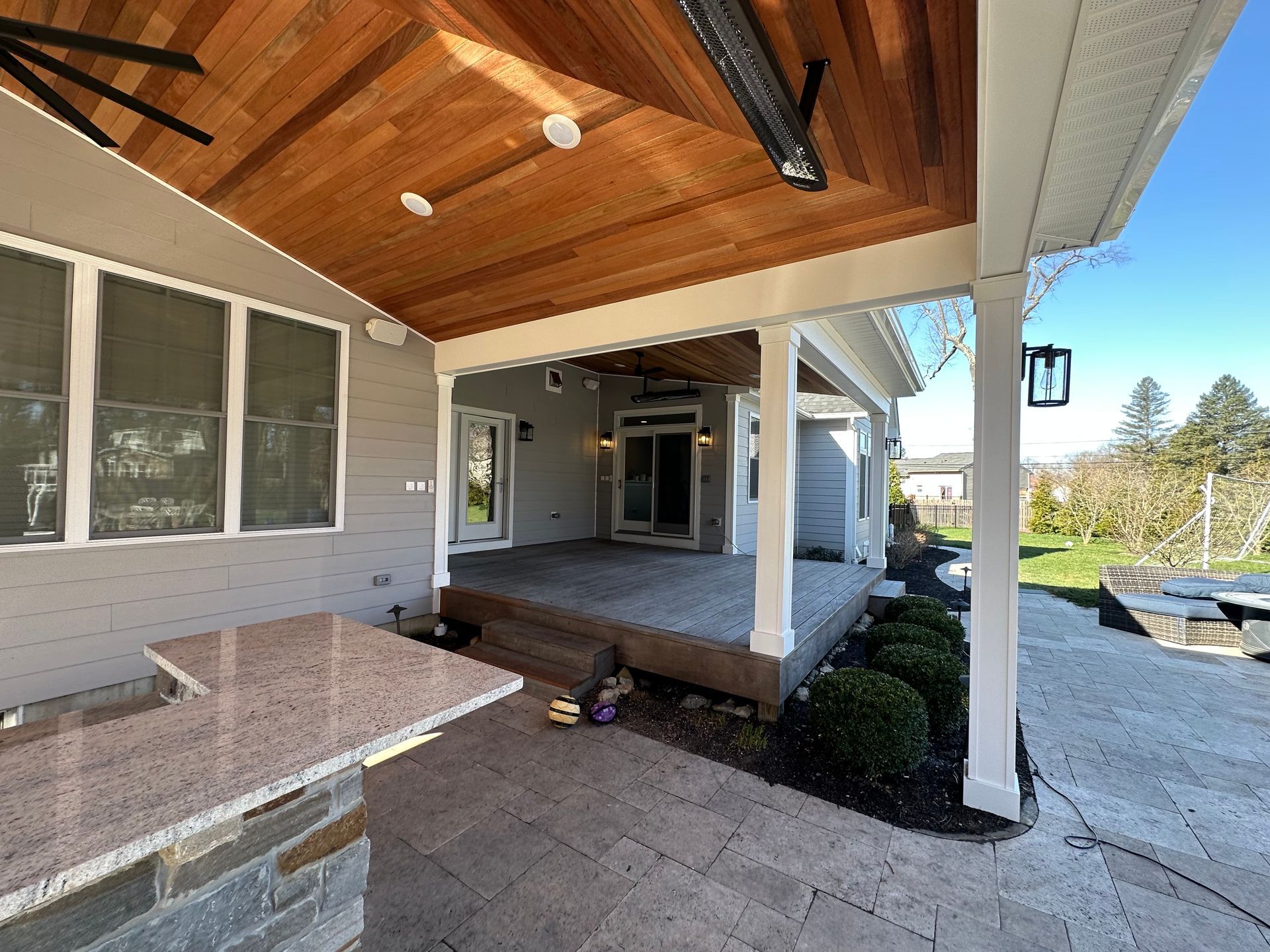 Covered front porch with wood ceiling, stone bench, white columns, and paved walkway outside a house.