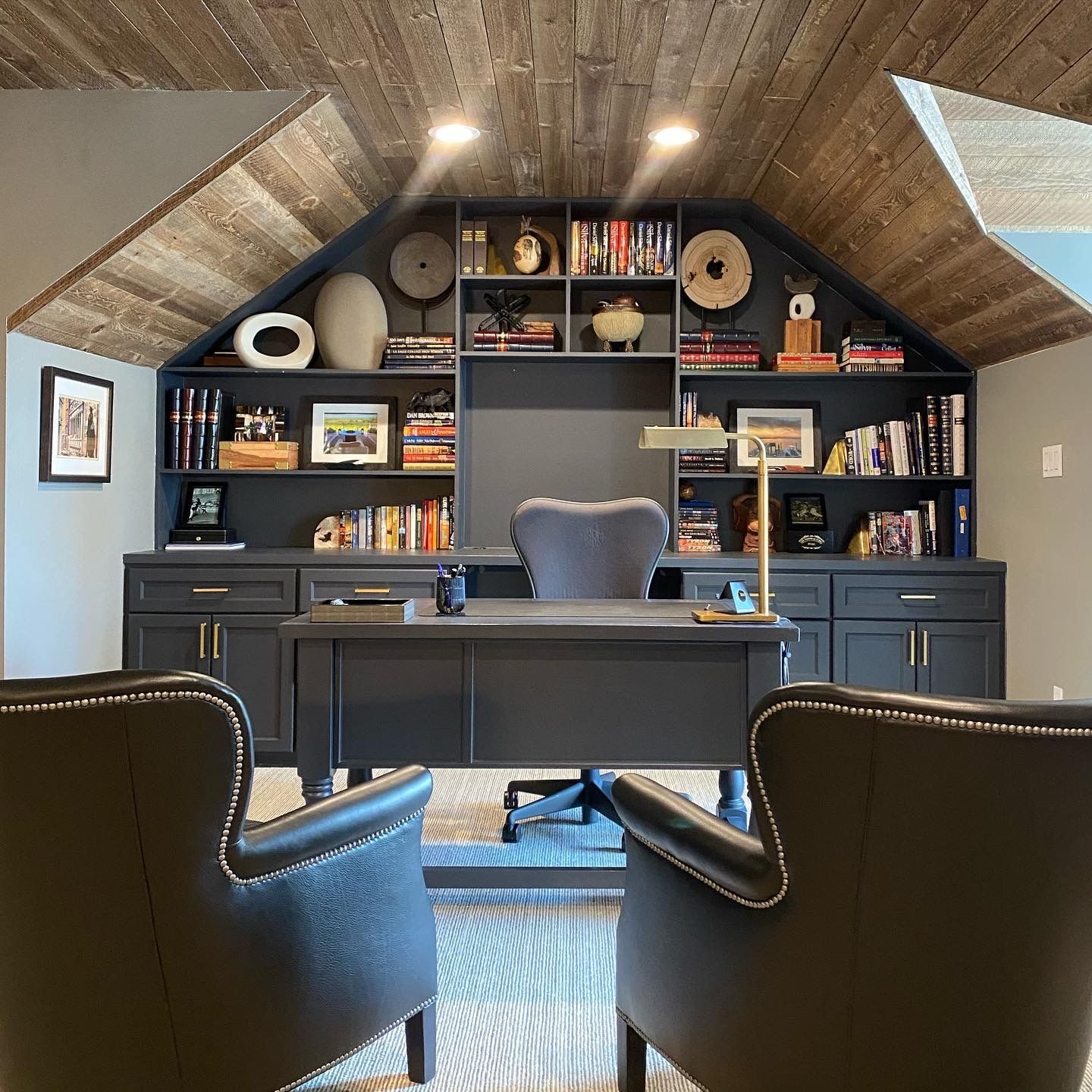 Modern home office with vaulted wood ceiling, dark built-in shelving, and a black desk with two chairs