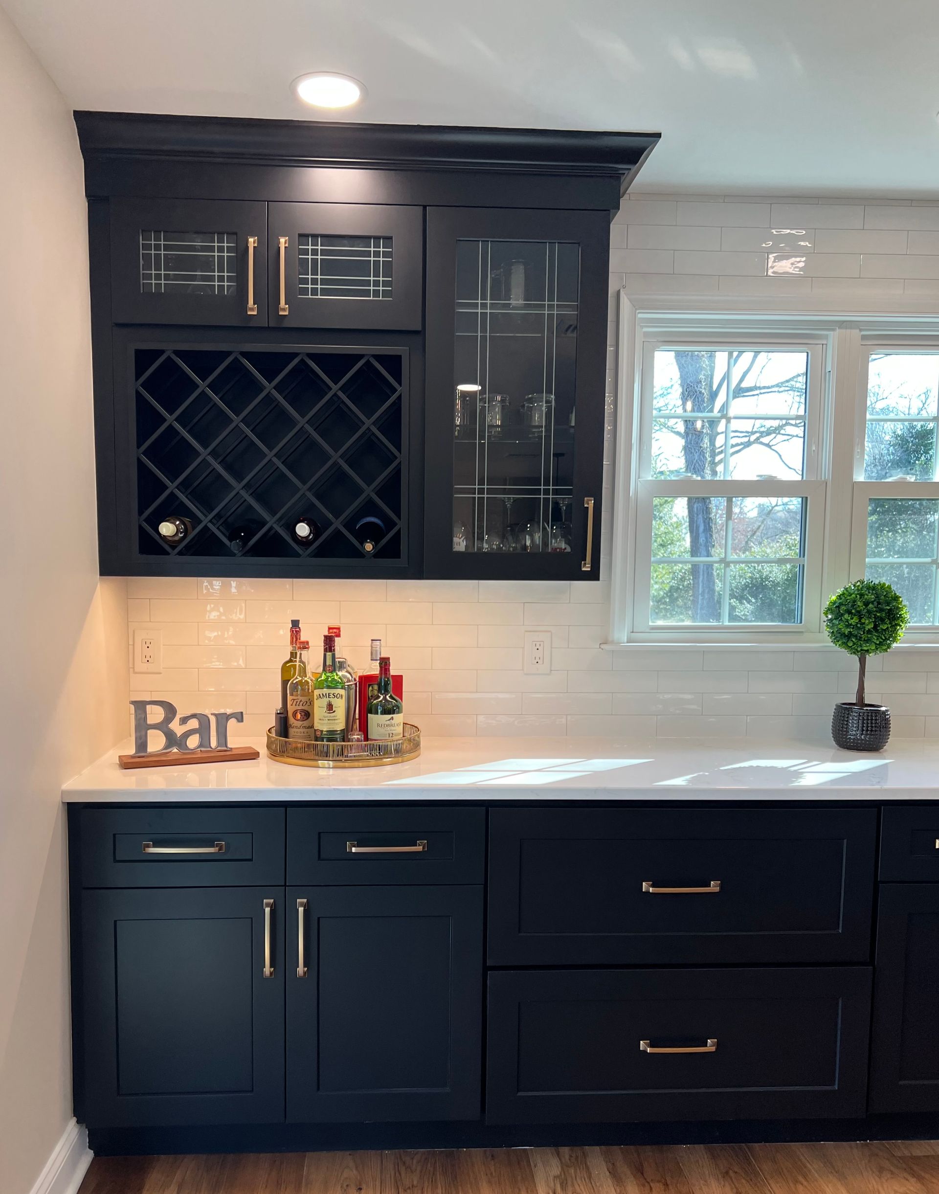 Black kitchen bar cabinet with wine rack, bottles, white countertop, and window above.
