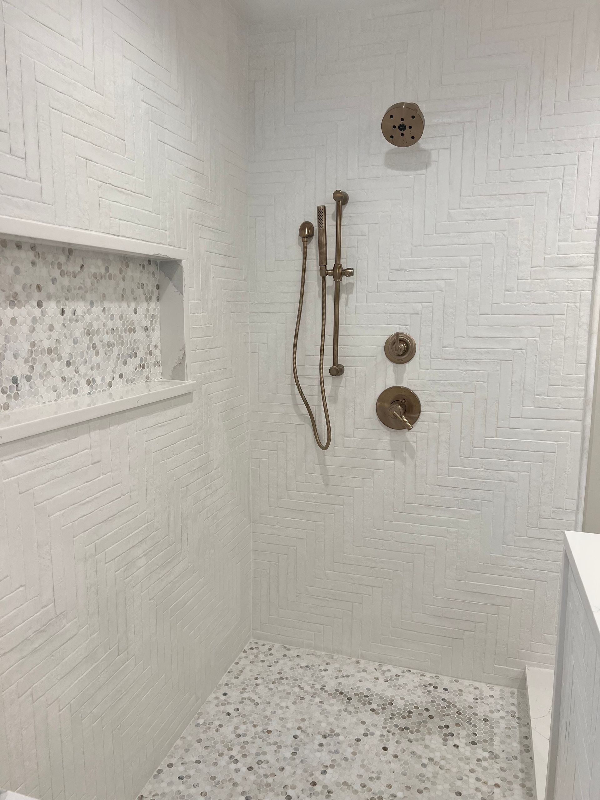 White tiled shower with built-in shelf, handheld showerhead, and two wall-mounted controls.