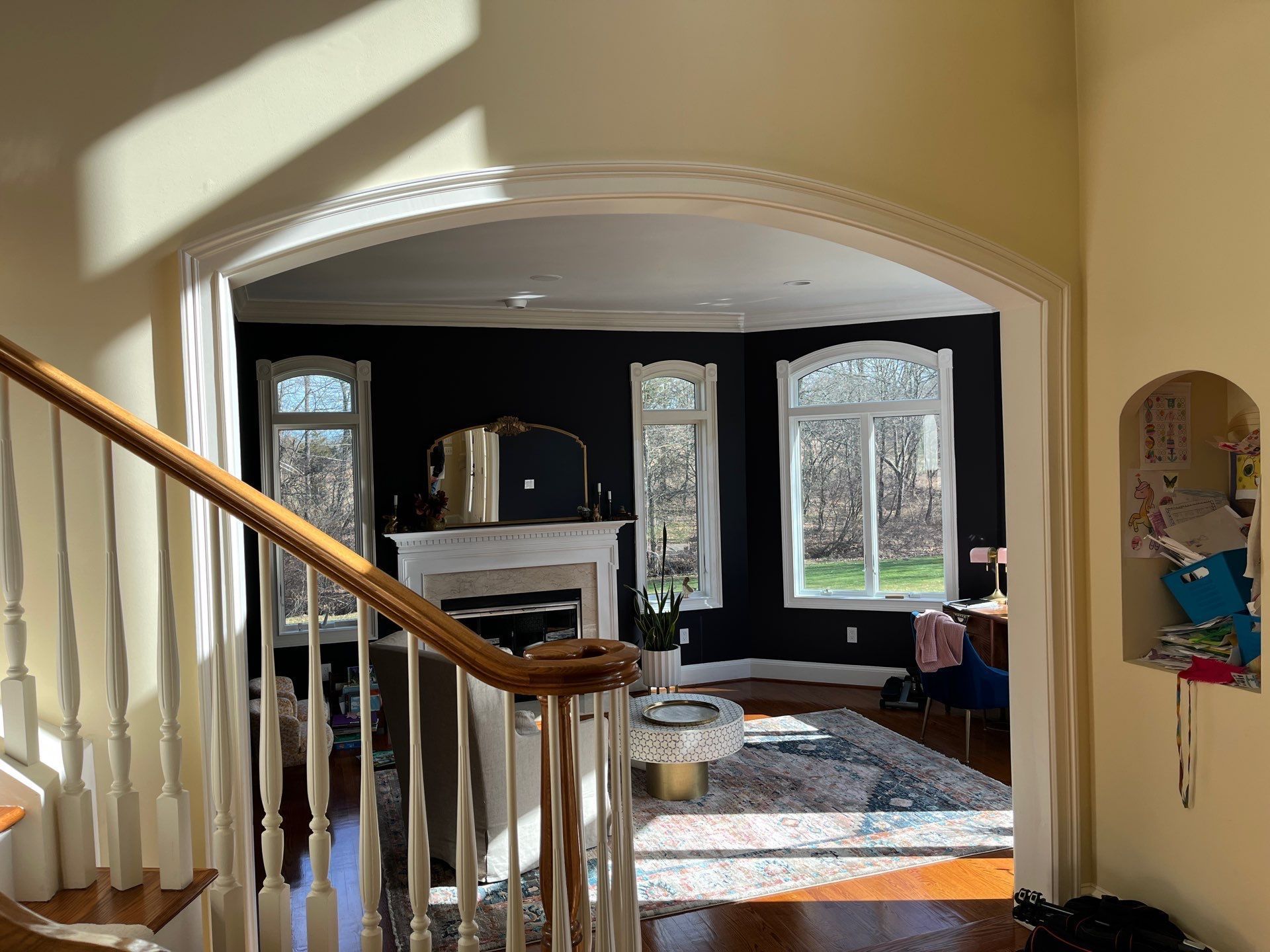 Staircase overlooks a bright living room with dark walls, white mantel, rug, and large windows.