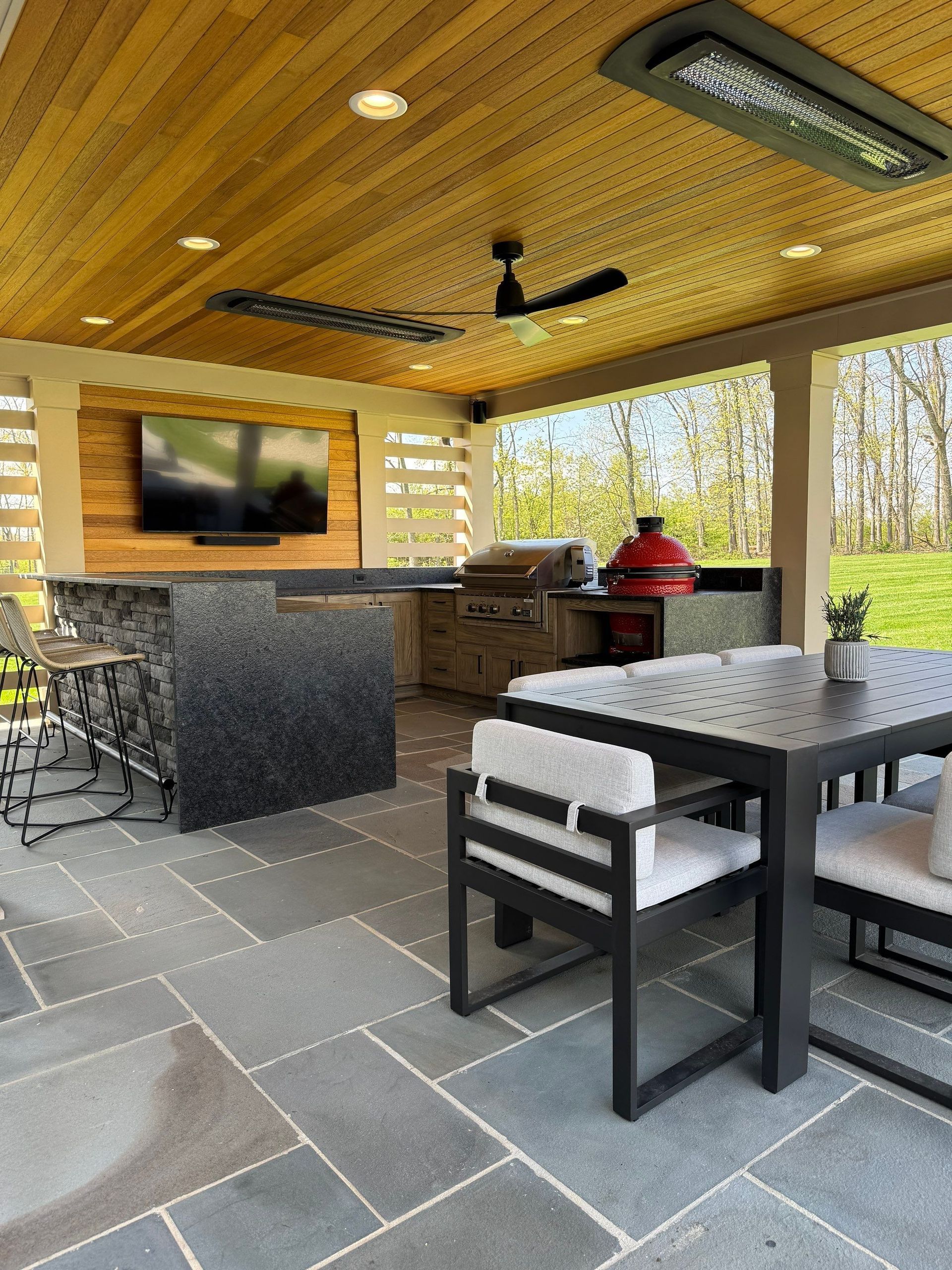 Covered patio with stone floor, dining table, grill, wall-mounted TV, and a person in a red shirt seated by the counter