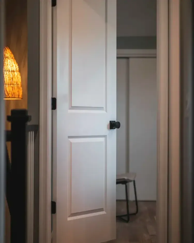 Partially open white door revealing a hallway with a small stool and warm lamp light on the left.