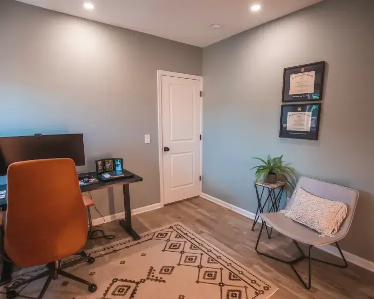Small home office with orange chair, desk, wall art, patterned rug, and a gray accent chair by a plant.