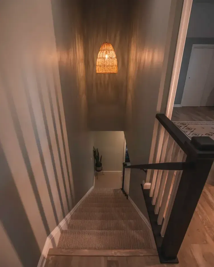 View down a carpeted staircase with a hanging woven pendant light and black railing in a dim hallway.