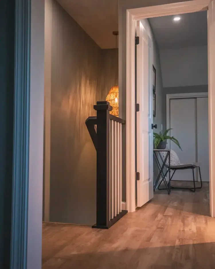 Hallway with warm wood floors, black staircase railing, open white door, and a chair with a plant in the room beyond