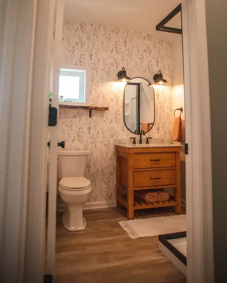 Small bathroom with toilet, wood vanity, round mirror, wall lights, and floral wallpaper.