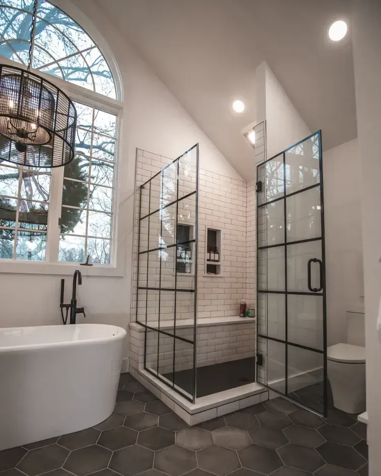 Modern bathroom with glass shower, white tub, black hex tile floor, and large arched window