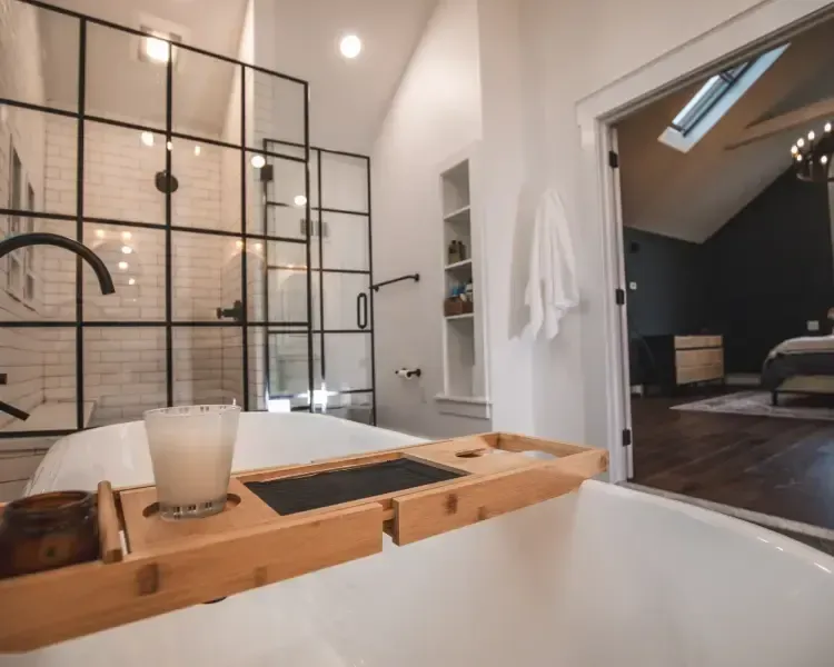 Modern bathroom with a freestanding tub, wooden bath tray, black metal shower divider, and open doorway to bedroom.