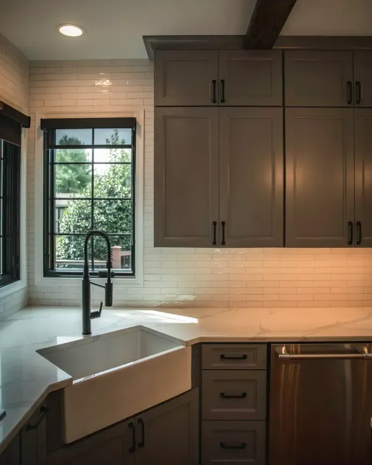 Modern kitchen sink under a window, with dark cabinets, white countertops, and black faucet.