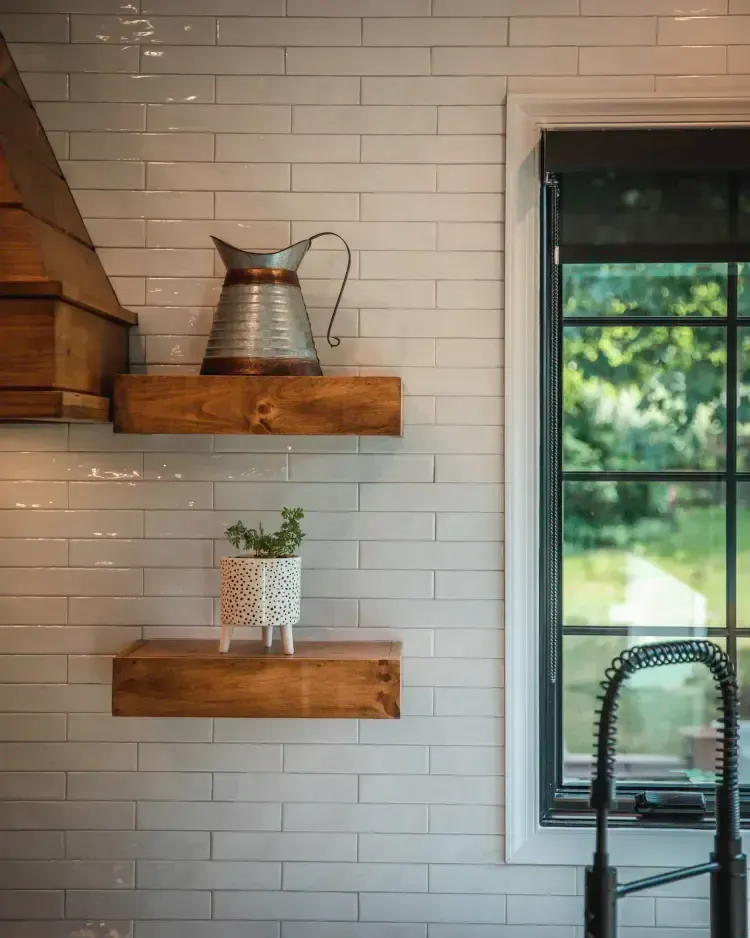 White subway tile kitchen wall with wood shelves, metal pitcher, small plant, and window by sink