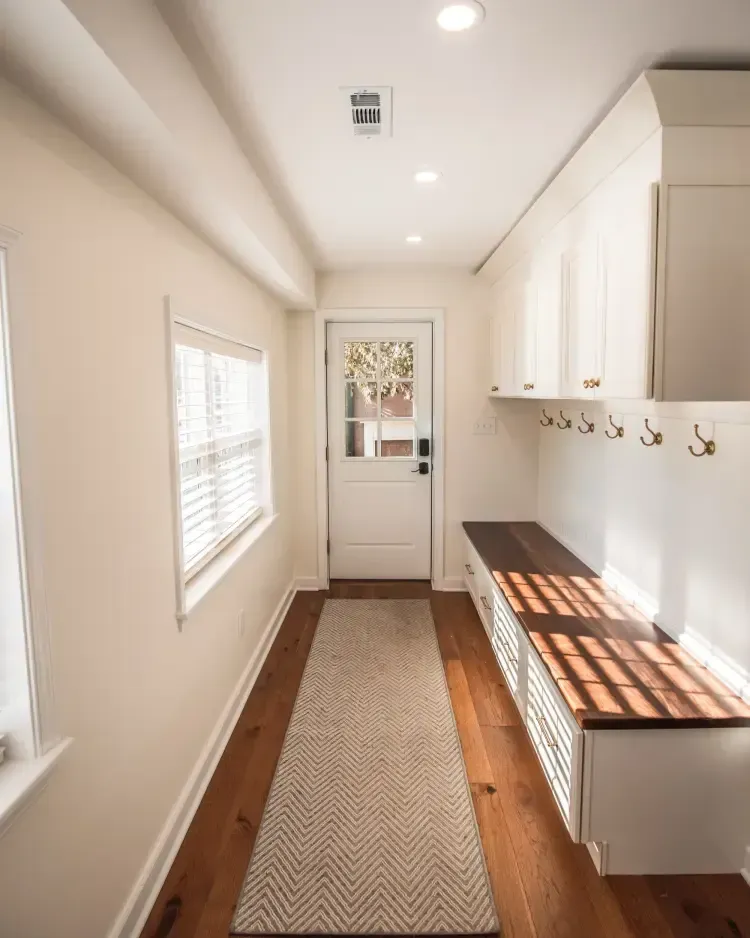 Narrow entry hall with white built-ins, wood bench, wall hooks, window, and a runner rug