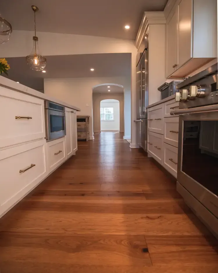 Modern kitchen with white cabinets, stainless steel appliances, and warm wooden floors leading to an arched hallway