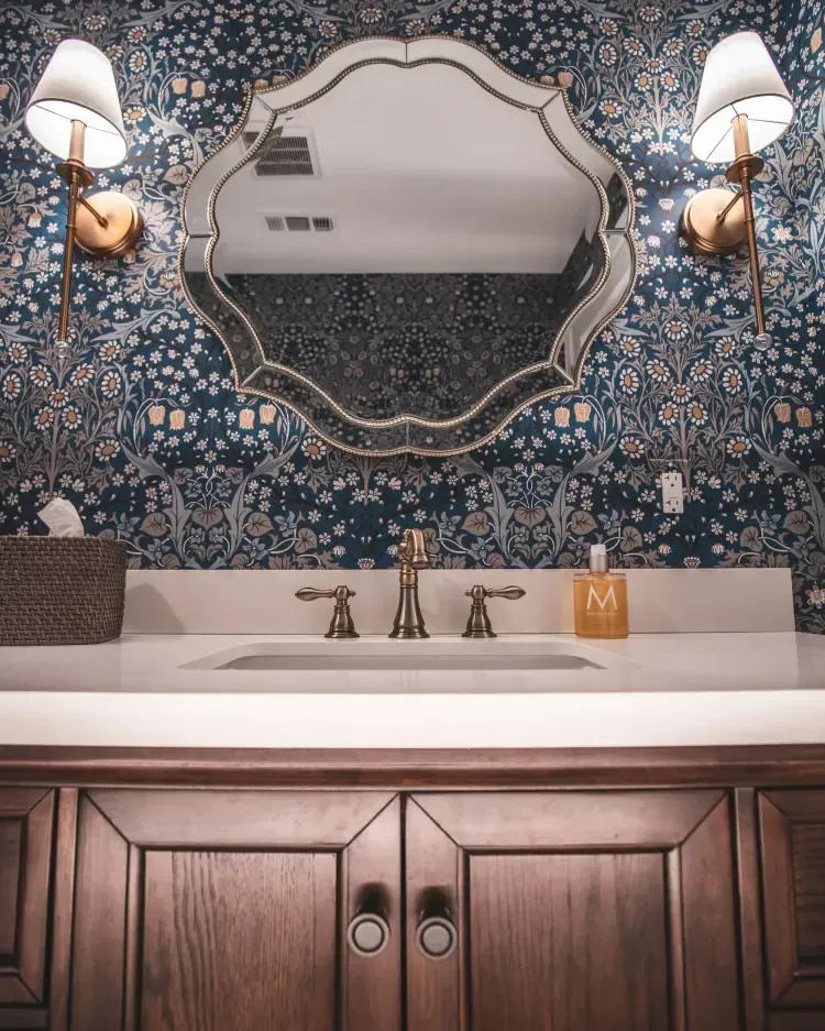 Elegant bathroom vanity with ornate mirror, blue patterned wallpaper, brass fixtures, and wood cabinet