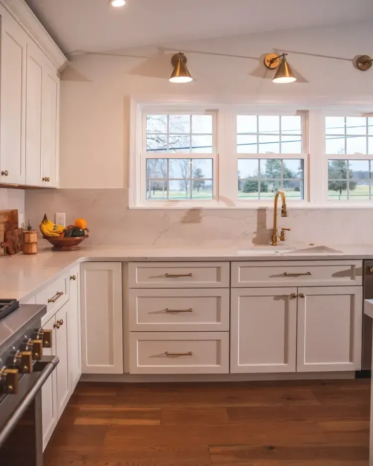 Bright white kitchen with farmhouse sink, marble counters, brass fixtures, and wood floors.