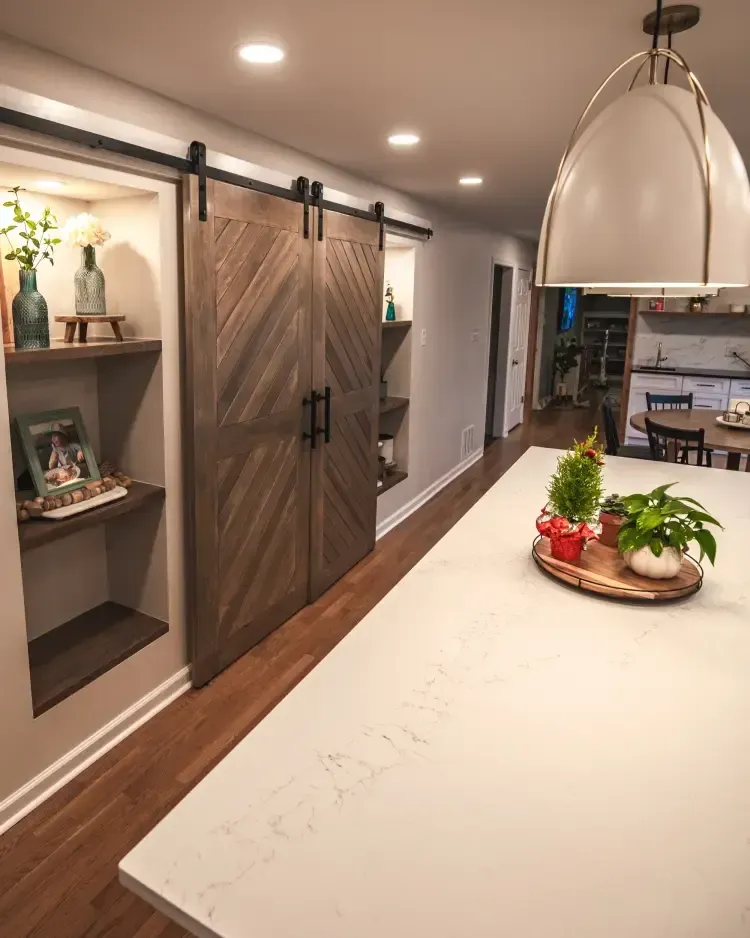Modern kitchen with a white island, pendant lights, barn-style pantry doors, and a tray of herbs and fruit.