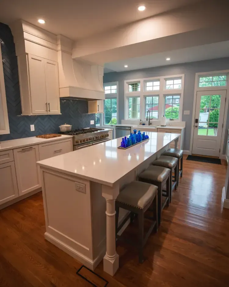 Bright modern kitchen with white island, gray stools, blue backsplash, and sunlit French doors.