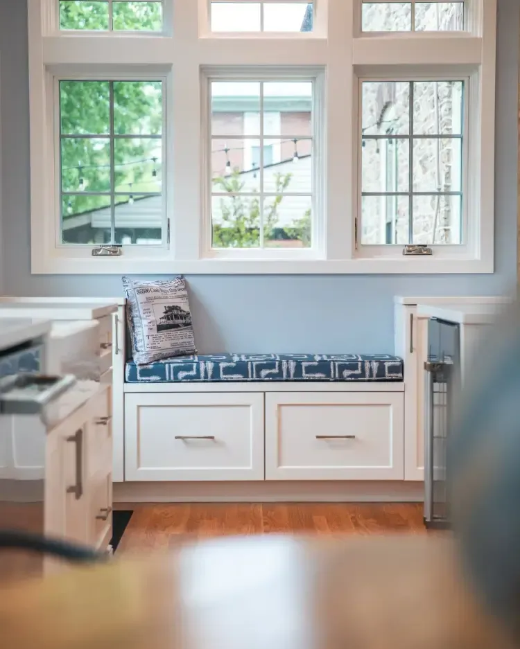 Built-in window seat with blue cushion and drawers beneath three large windows in a bright room