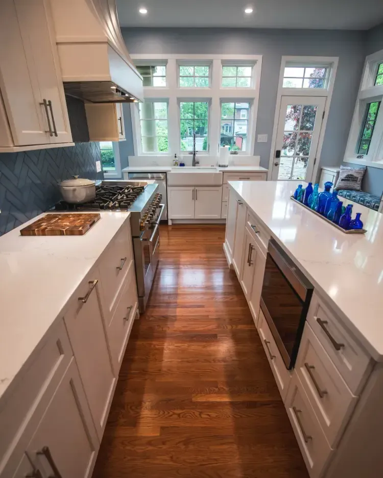 Bright modern galley kitchen with white cabinets, wooden floors, and large windows at the far end