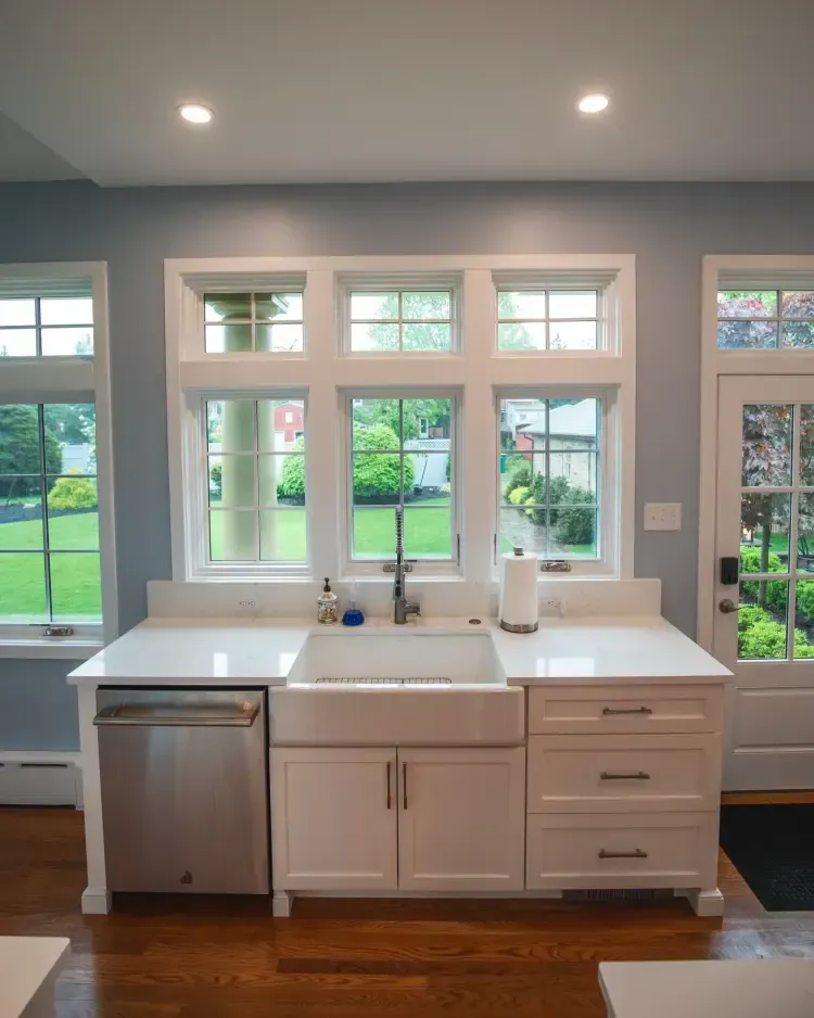 Bright modern kitchen sink under windows, with white cabinets, stainless dishwasher, and green backyard views.
